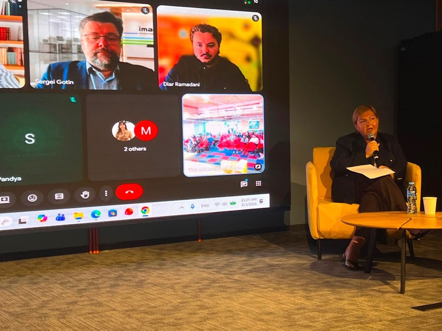 The panel moderator with a microphone; on the screen, the panellists joining remotely and a view of the hall.